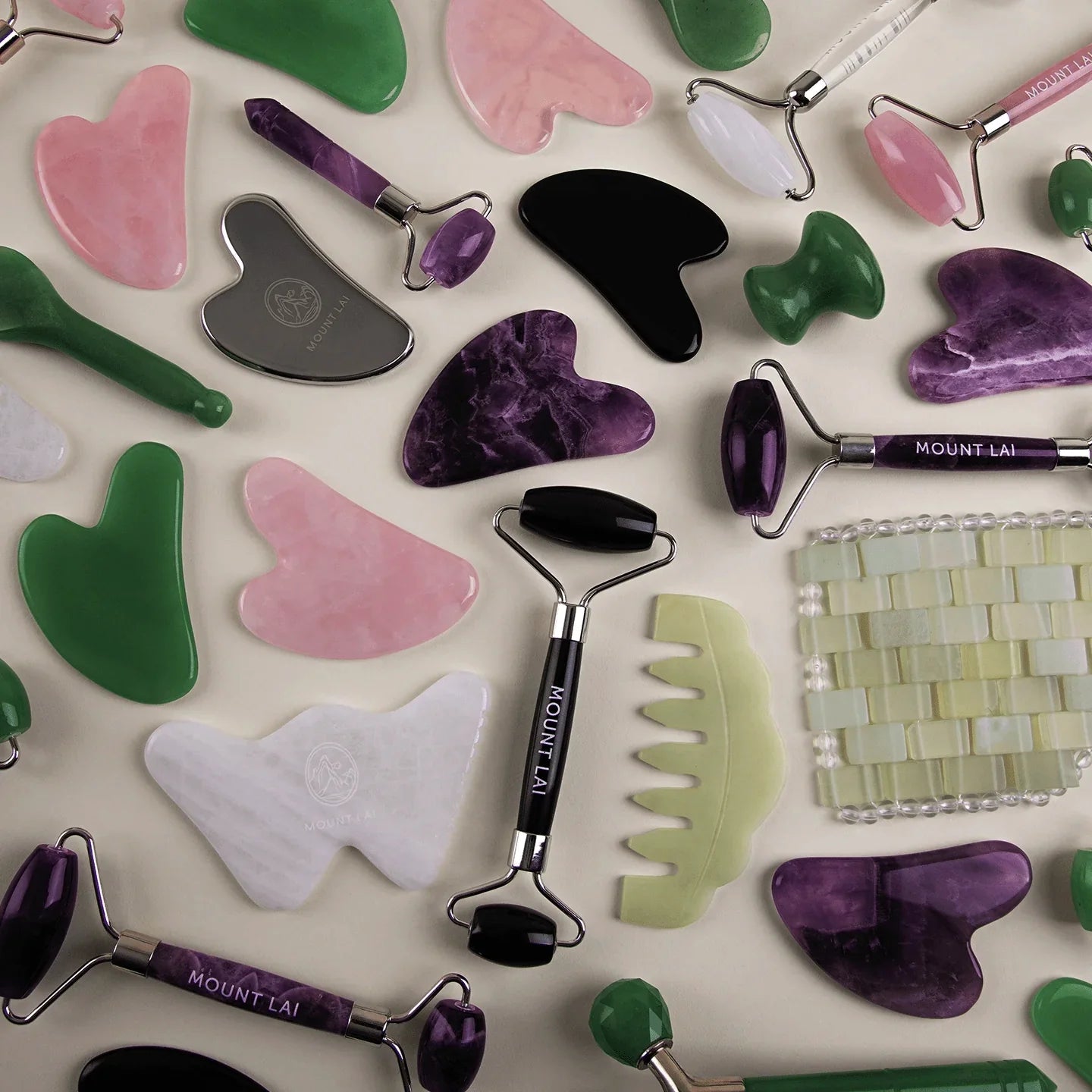Assorted Mount Lai facial tools, including jade rollers and gua sha, in green, pink, white, black, and purple stones.