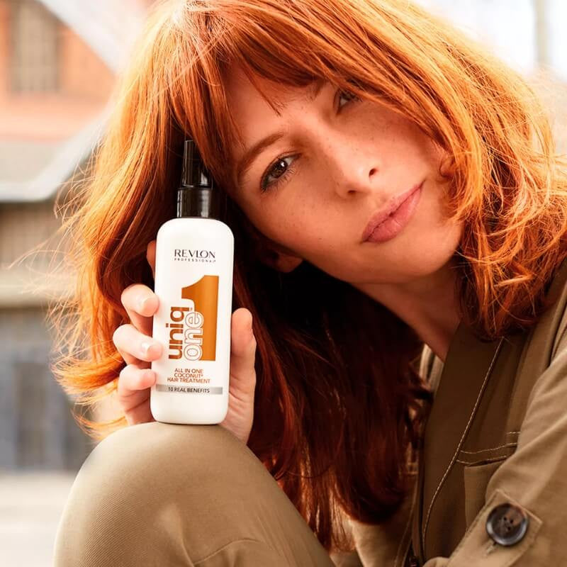 Woman with red hair holding Revlon Uniq One Coco Leave-in, promoting its effectiveness for all hair types and visibly healthier hair.