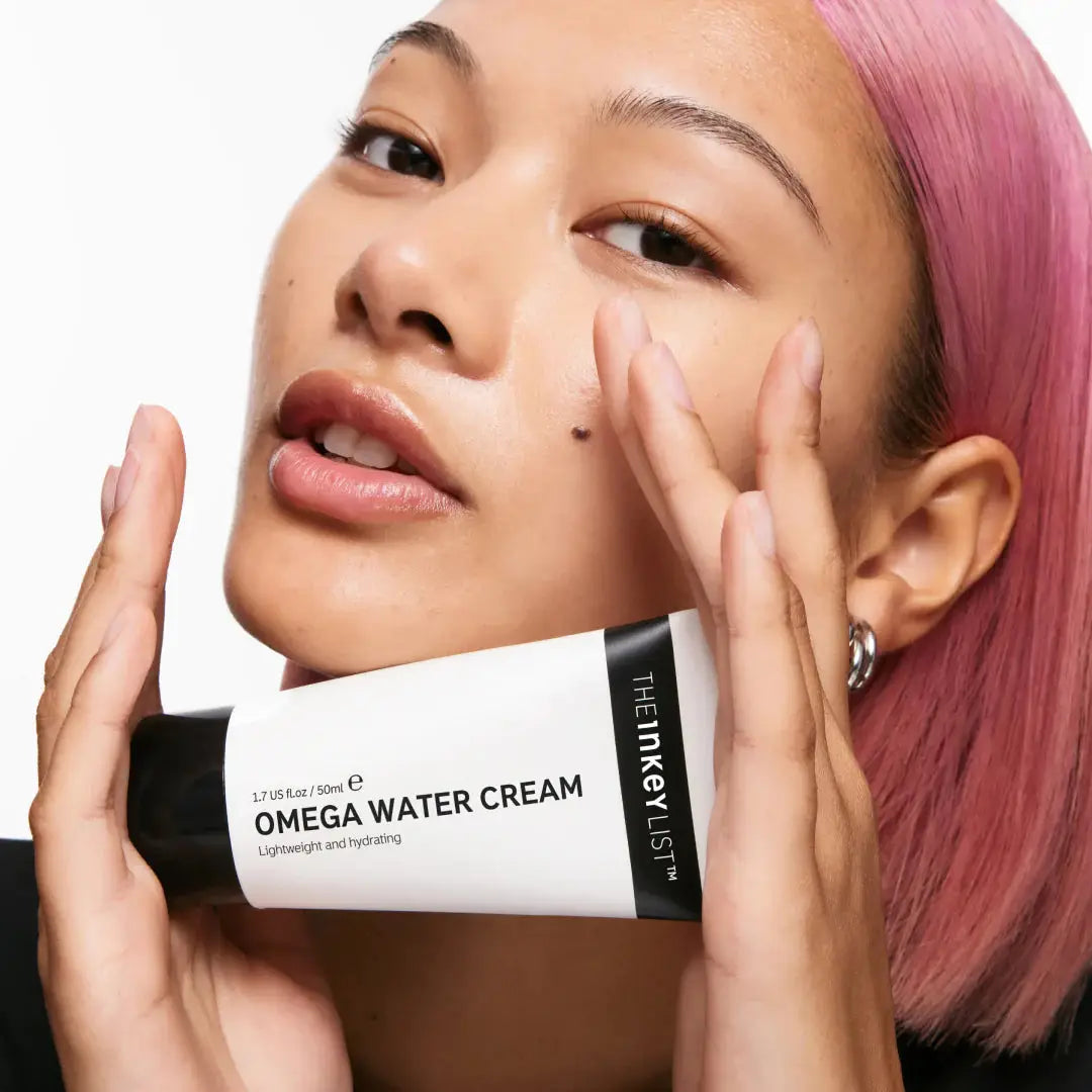 A model with pink hair holding The Inkey List Omega Water Cream, emphasizing its lightweight, hydrating formula for smooth and healthy-looking skin.