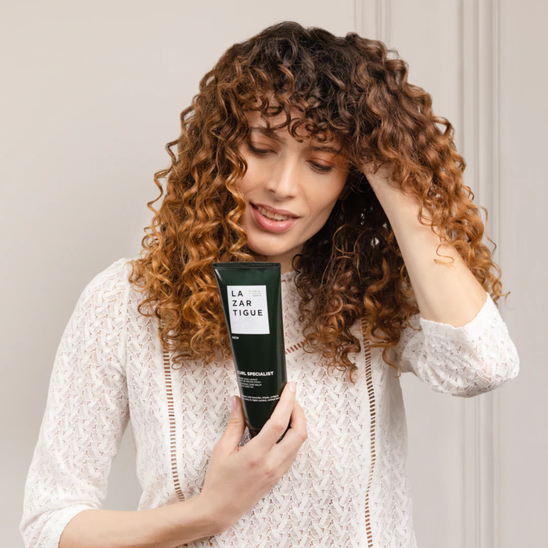 Woman with defined curly hair holding Lazartigue Curl Balm, ideal for frizz control and curl enhancement in natural hair care routines.