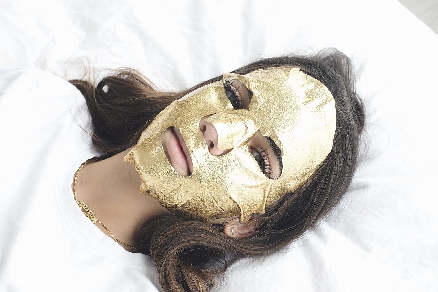 Woman lying down wearing Masque Bar Gold Foil Sheet Mask, a facial treatment that hydrates and strengthens the skin barrier.