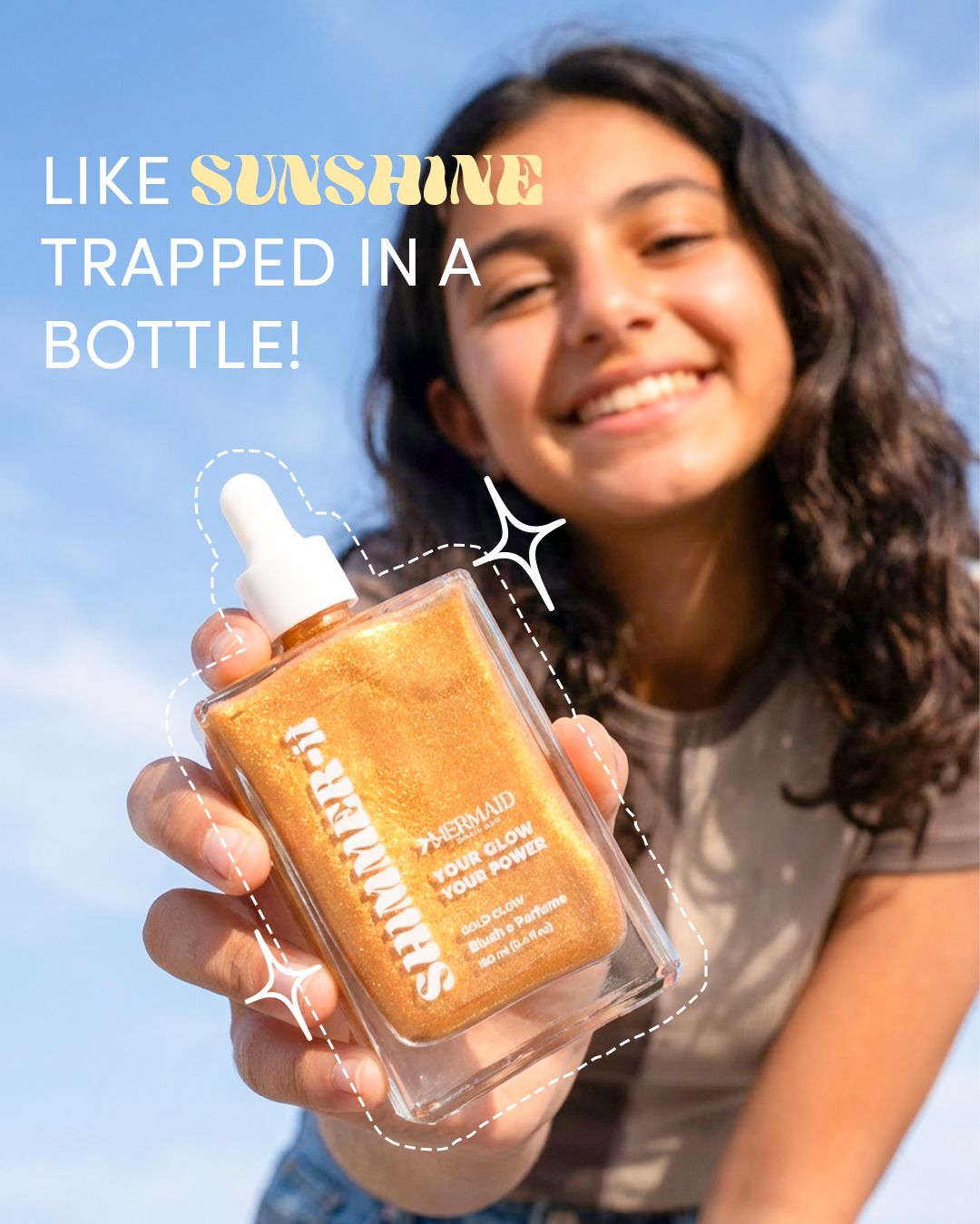 Smiling woman holding Mermaid Gold Shimmering Oil against blue sky. Lightweight shimmering body oil for glowing skin with Blushe Perfume.