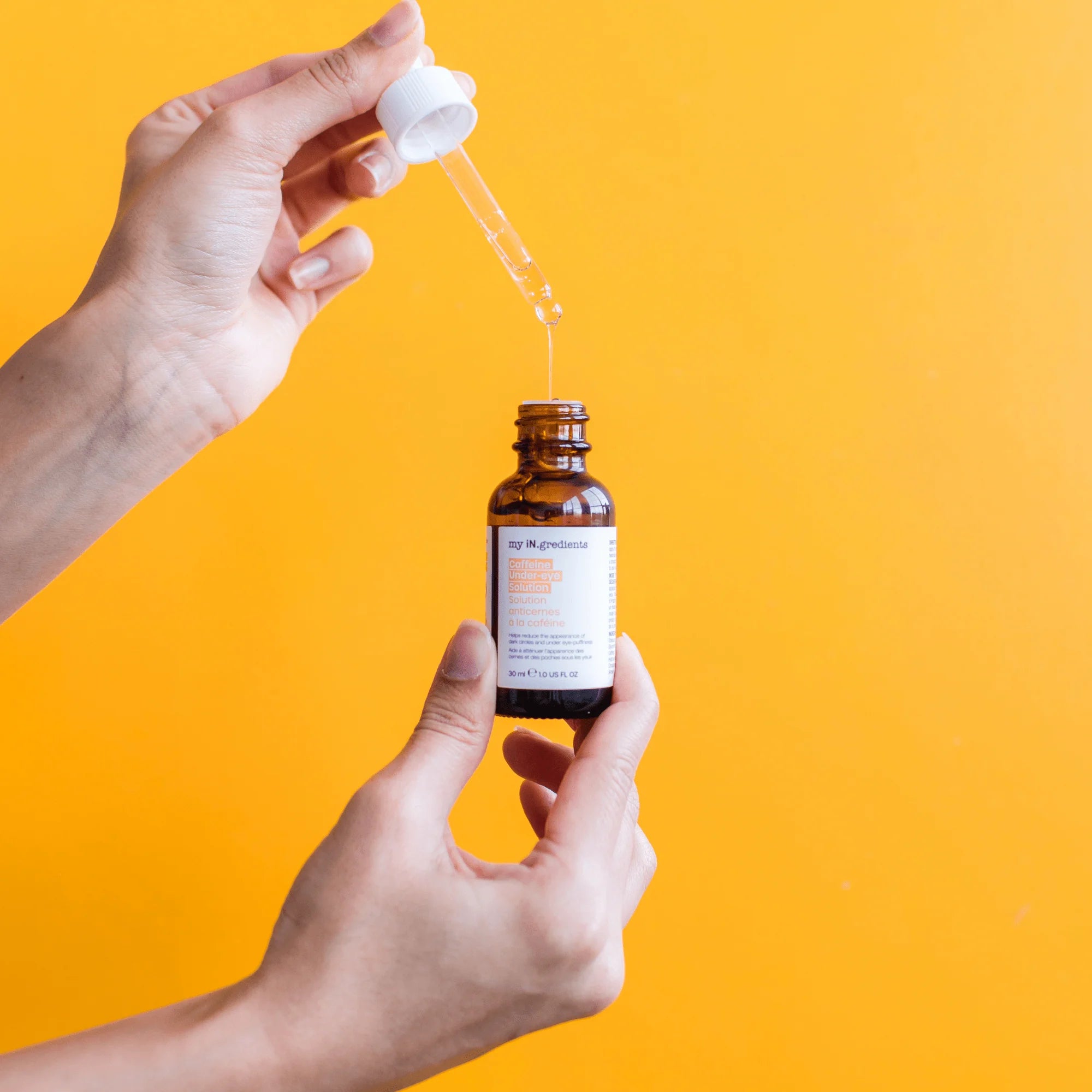 Hands holding iN.gredients Caffeine Under-Eye Solution with dropper, showing lightweight serum texture for brightening under-eye area.