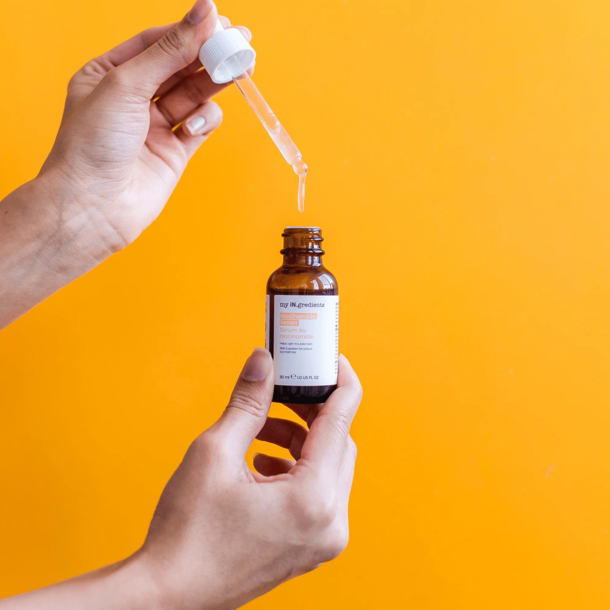 Hand holding My iN.gredients Niacinamide Serum 30 ml dropper against a bright orange background, perfect for smoothing and toning skin.
