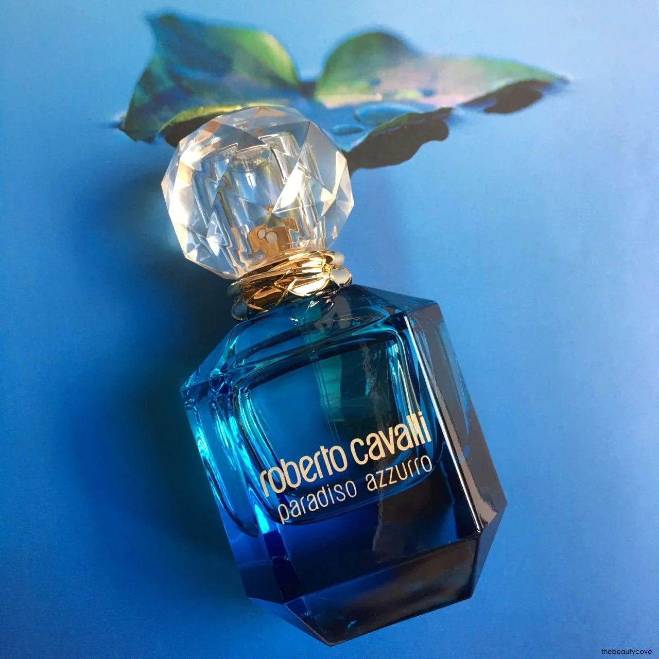 Roberto Cavalli Paradiso Azzurro Eau De Parfum 75 ml displayed on a blue background, showcasing its elegant crystal-cut cap and vibrant blue glass bottle.