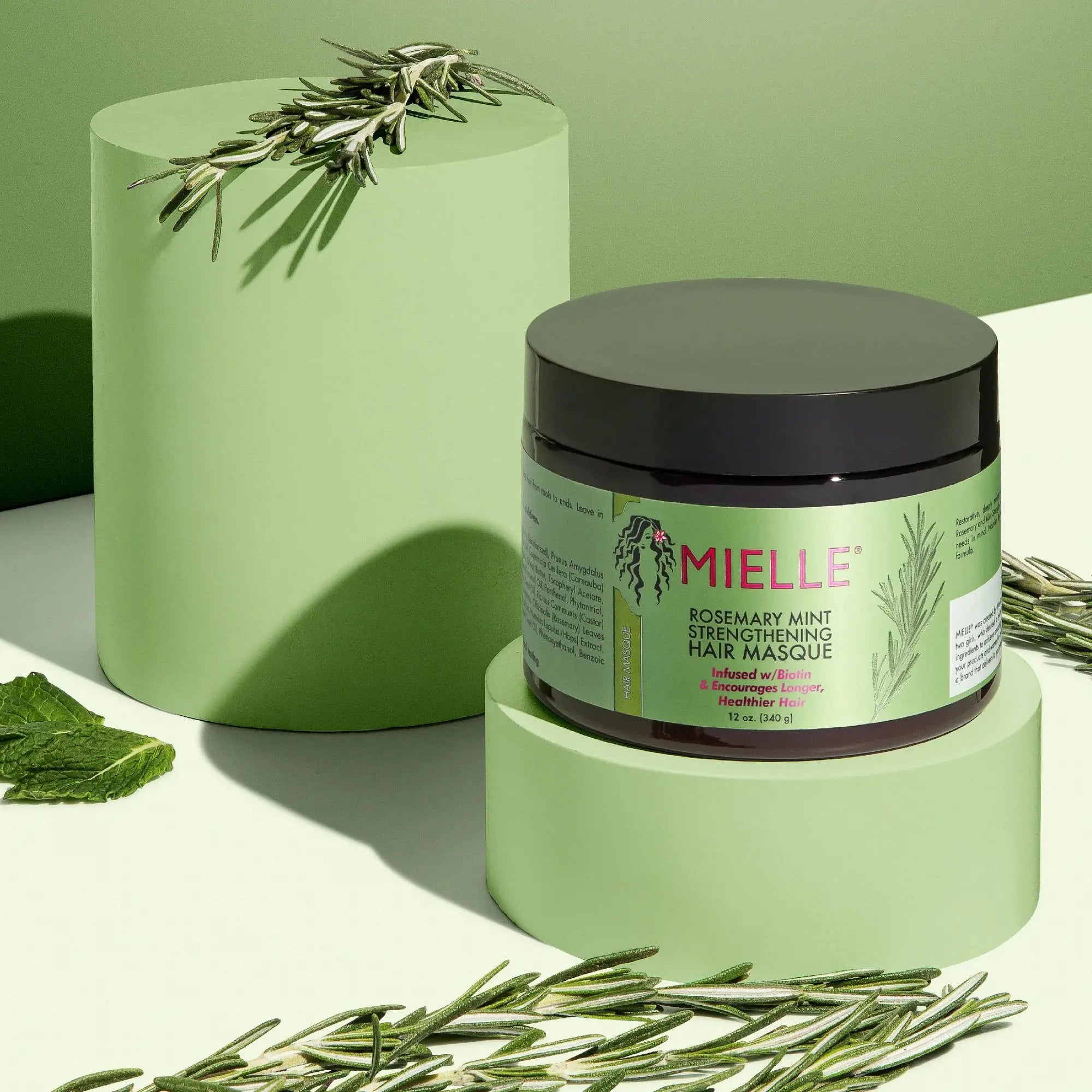 Mielle Rosemary Mint Strengthening Hair Masque 340 G displayed on green platforms with rosemary and mint leaves, highlighting its natural ingredients.