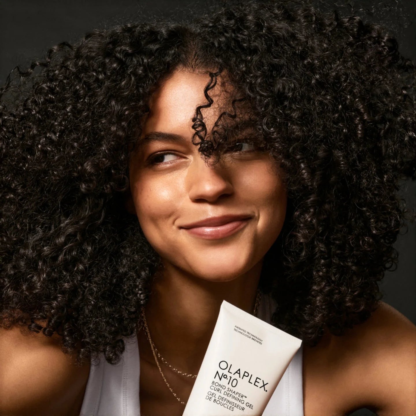 Smiling woman with defined, voluminous curls holding Olaplex No.10 Curl Defining Gel, highlighting its frizz control and curl-enhancing benefits.